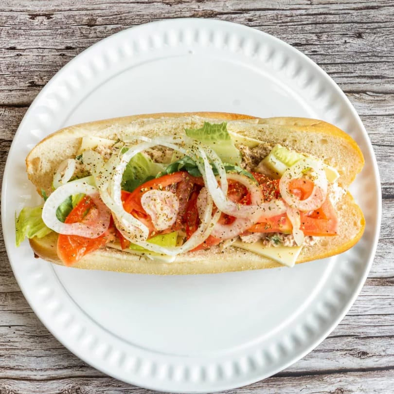 A tuna sub sandwich with lettuce, tomatoes, onions, and cheese on a white plate.