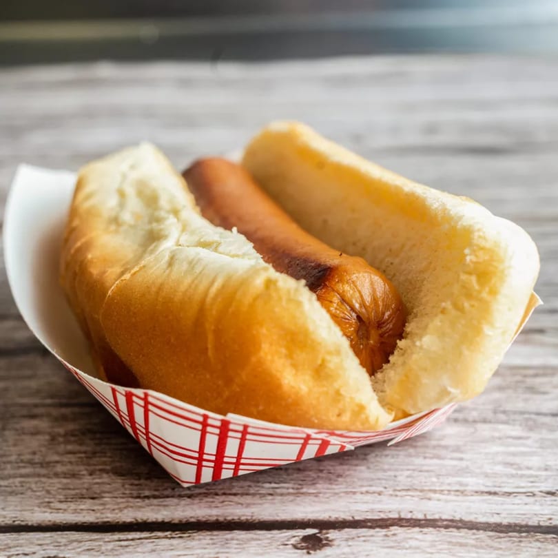 Grilled hot dog in a white bun, served in a red and white checkered paper tray.