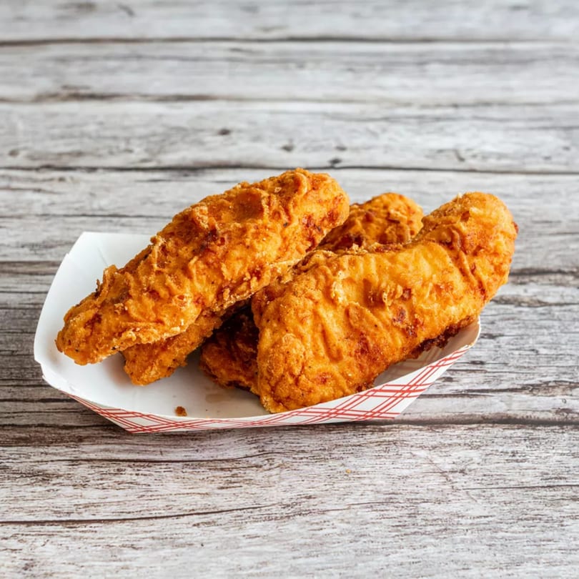 Golden-brown fried chicken tenders in a white paper tray with red checkered trim on a wooden surface.